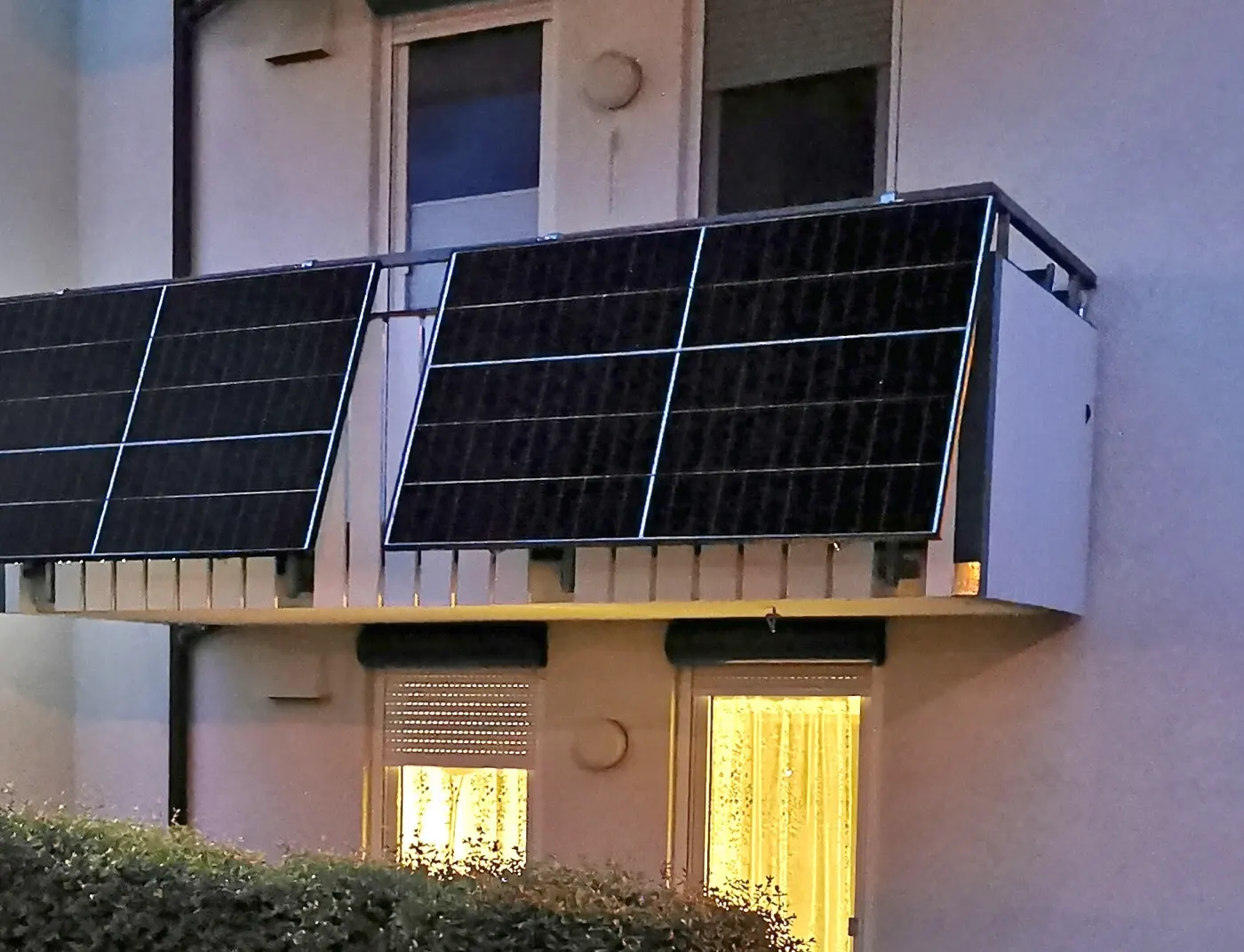 Four solar panels clipped to the railing of a ground-floor apartment balcony, at dusk.