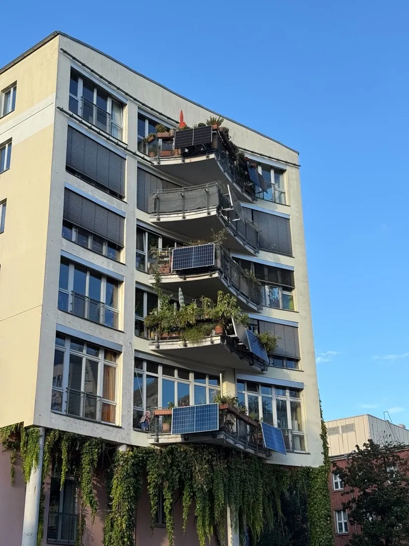A tall apartment building with solar panels on several balconies, some flat against the railing and some angled outward.