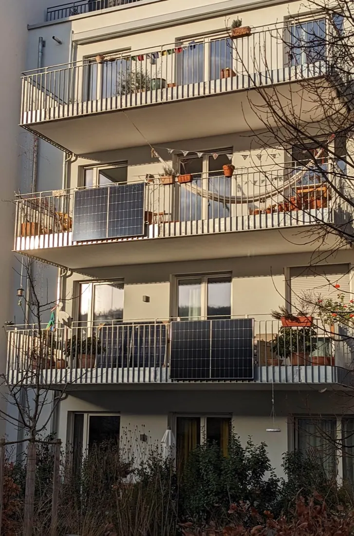 Two balconies of an apartment building, each with two black solar panels hung on its railing, potted plants behind.