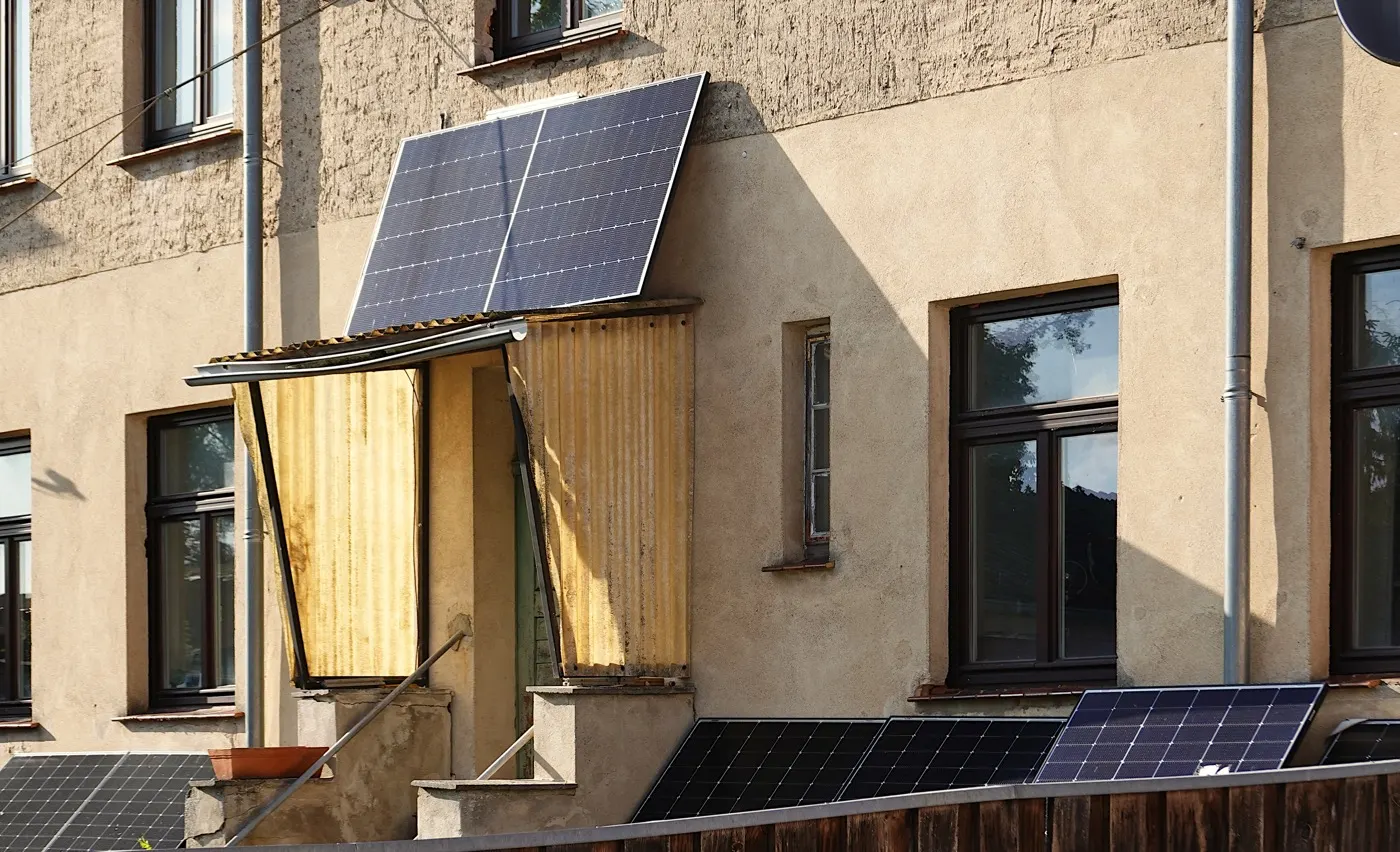 An old apartment building with two solar panels mounted on a porch roof and more panels leaning at an angle along a garden fence.