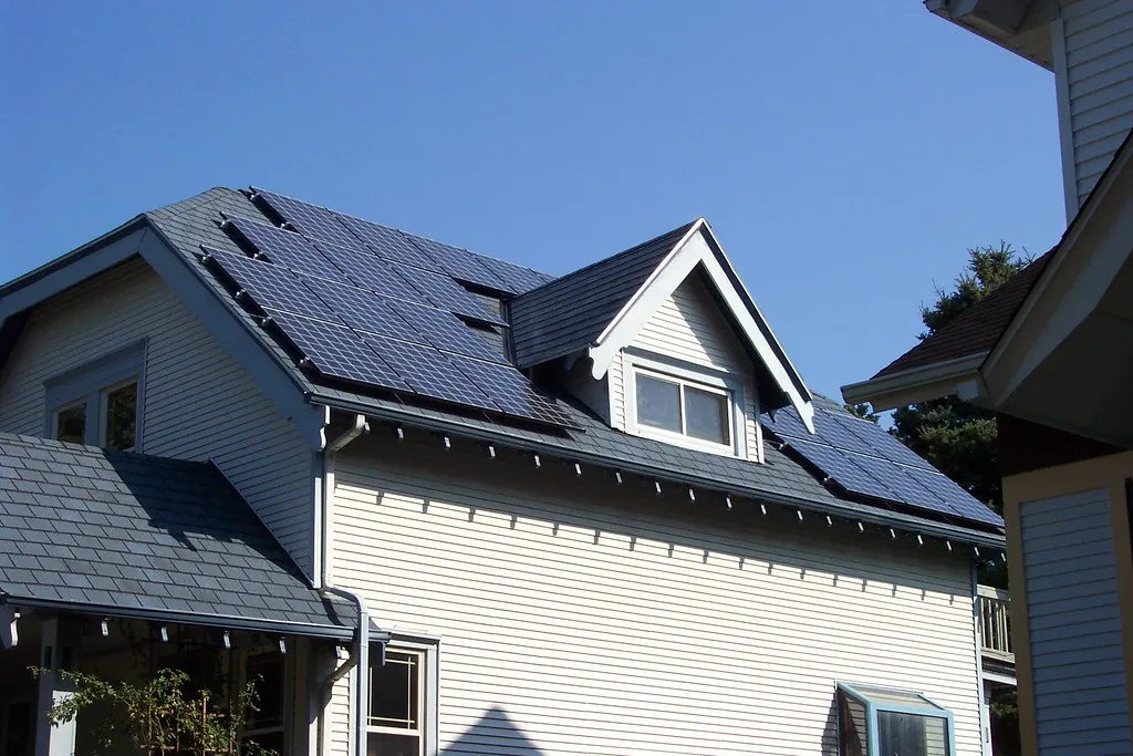 A two-story house with solar panels across its roof, seen from the street on a sunny day.