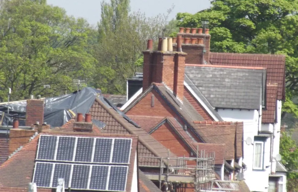 Solar panels on a tiled roof, with brick chimneys and tall trees on the houses beside them.