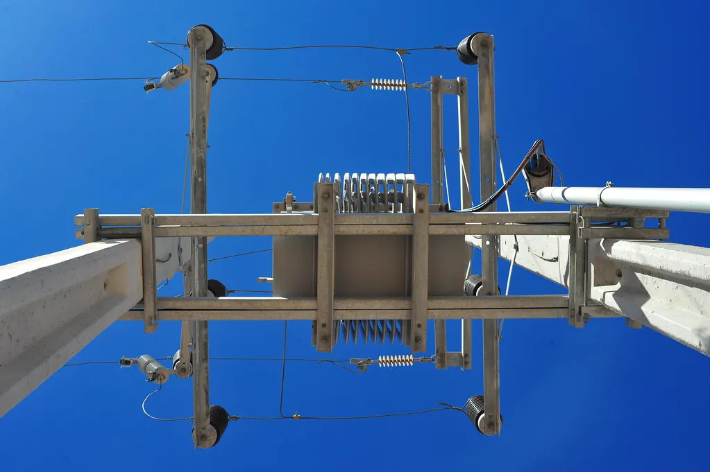 Looking straight up a concrete power pole at utility equipment, insulators, and wires against a blue sky.